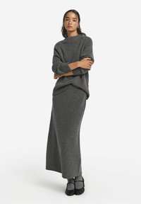 Grå stickad tröja med hög halsringning, matchad med en lång kjol. Texturerat material och avslappnad passform. Svarta skor fulländar outfiten.