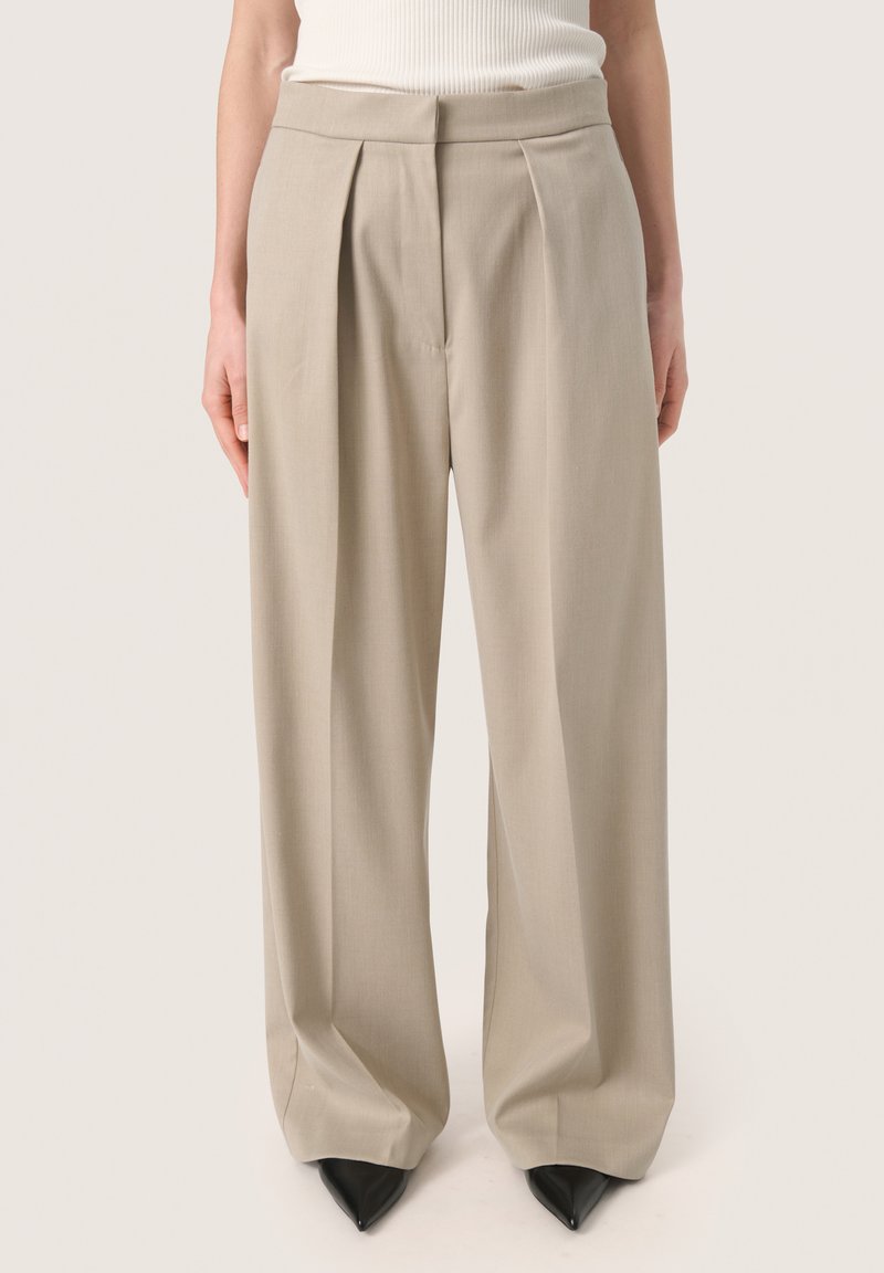 Pantalons larges beiges en tissu lisse, avec des plis à l'avant et une taille haute. Associés à des talons aiguilles noirs.