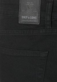Gros plan sur la poche arrière d'un jean en denim noir avec un patch en cuir cousu portant l'inscription "Only & Sons, Passionate Jeans Makers."