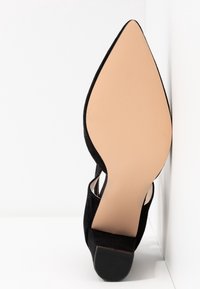 Chaussure noire en daim à bout pointu avec semelle beige et petit talon, présentée debout sur fond blanc.