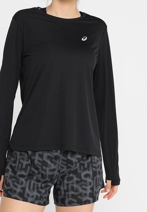 Kobieta w czarnej, długorękawowej koszulce Asics i czarnych, wzorzystych szortach, stoi na tle jednolitego białego tła.