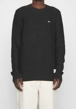 Mann trägt einen schwarzen Strickpullover mit dezentem Logo auf der Brust und einem Etikett am Saum, kombiniert mit lockeren, cremefarbenen Hosen vor einem schlichten Hintergrund.