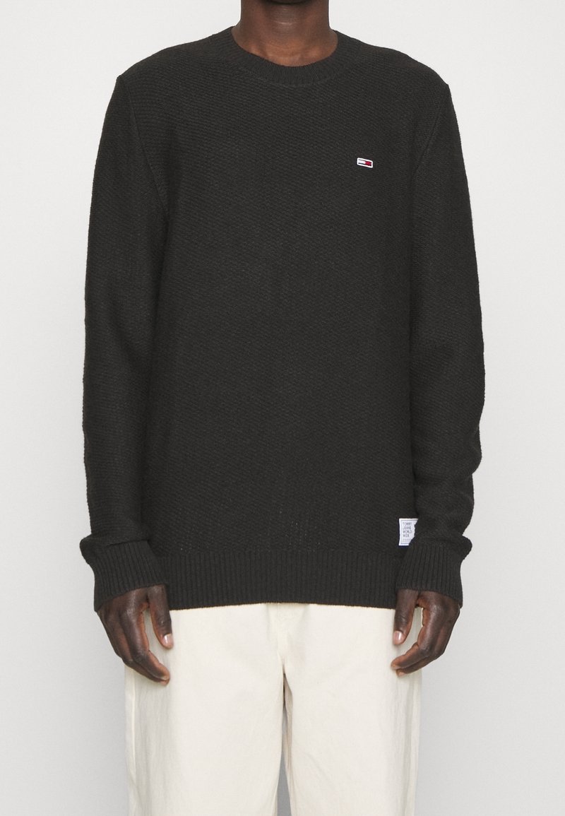 Homme portant un pull en tricot noir avec un logo subtil sur la poitrine et une étiquette sur l'ourlet, associé à un pantalon ample blanc cassé sur fond uni.