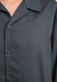 Mörkgrå skjorta med knappar och en mjuk textur. Har en spetsig krage och en broderad "AIR JORDAN"-logotyp i grönt på bröstet.