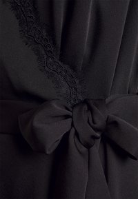 Svart plagg med mjuk textur, prydd med delikata volanger längs kanten och en knuten rosett vid midjan för en åtsittande look.
