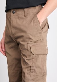 Pantalones cargo marrones con una textura suave, que cuentan con bolsillos laterales y un cierre de botón. El material parece duradero y tiene un sutil patrón en cuadros.