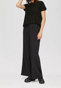 Svart t-shirt med upphöjd logotyp, ihop med lösa, mörka vida byxor. Texturen verkar mjuk, med en avslappnad passform. Svarta skor fulländar looken.