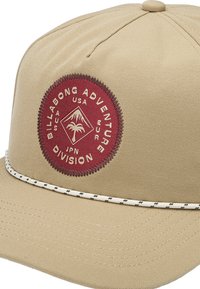 Casquette en tissu beige avec un patch rond bordeaux comportant un texte blanc et un graphique de montagne, accentuée par une bande texturée blanche le long de la visière.