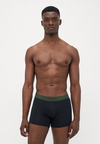Boxer briefs pretos com uma faixa verde da Levi's, apresentando um ajuste justo e uma textura suave. O modelo está em pé com uma expressão neutra.
