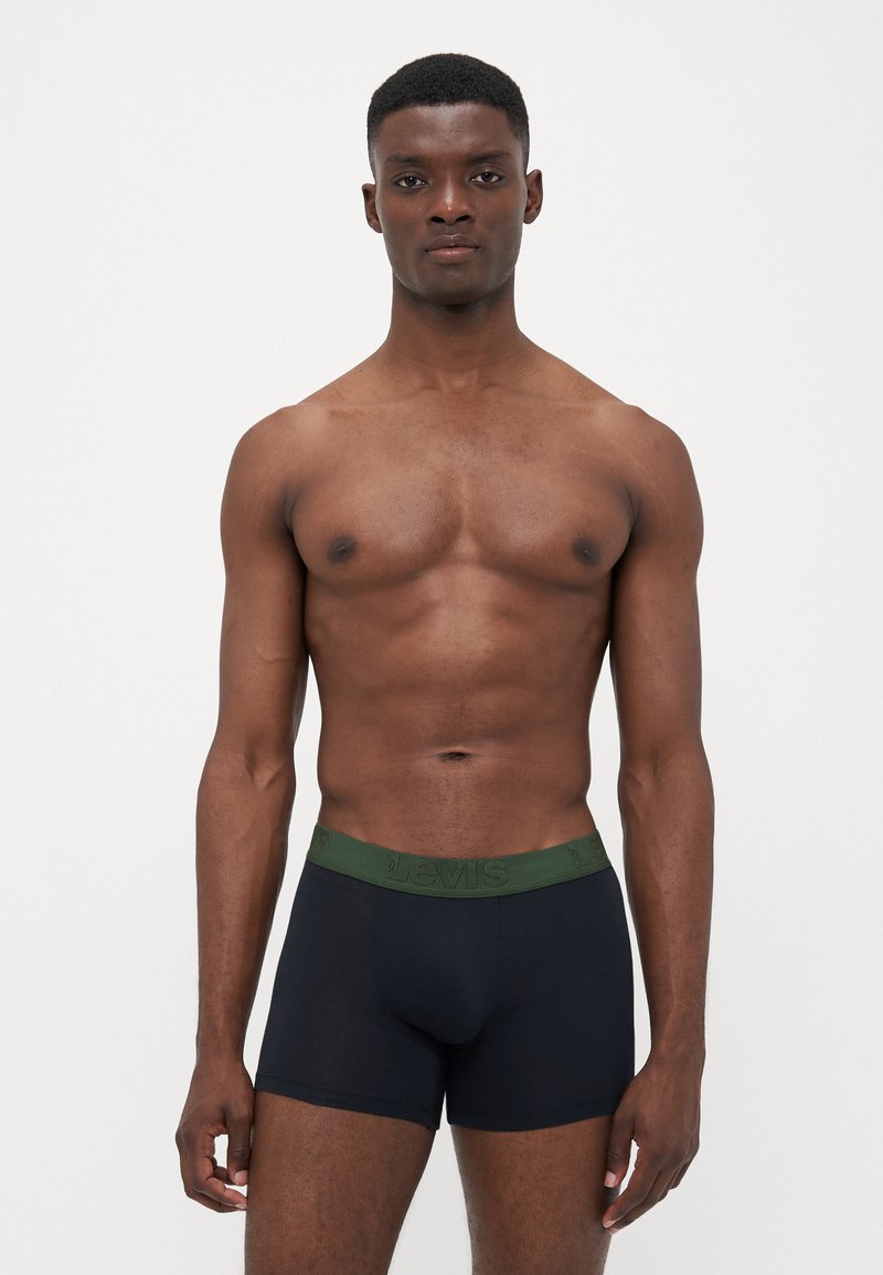 Boxer briefs pretos com uma faixa verde da Levi's, apresentando um ajuste justo e uma textura suave. O modelo está em pé com uma expressão neutra.