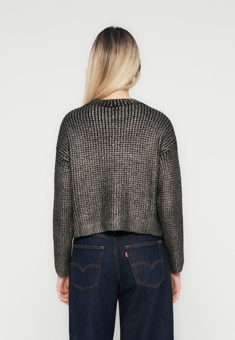 Donna con lunghi capelli biondi, che indossa un maglione grigio di maglia testurizzata e jeans blu scuro, in piedi di fronte a un muro bianco semplice.