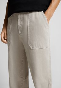 Henkilö, joka on pukeutunut vaaleanbeigeihin väljät housuihin, joissa on suuret etupocketit, ja mustaan puseroon, yksi käsi taskussa, tasaisella taustalla.