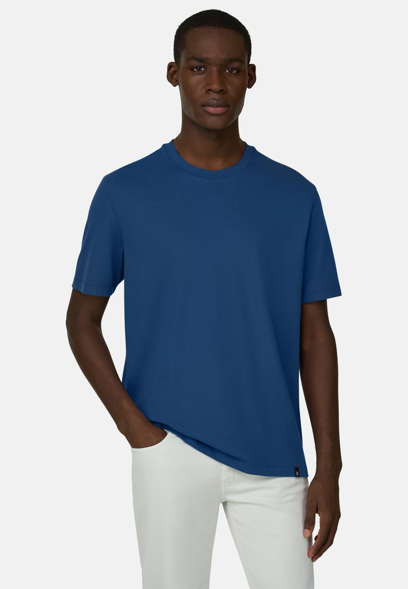 Homme portant un t-shirt bleu uni à manches courtes et un pantalon blanc, debout avec une main dans la poche devant un fond uni.
