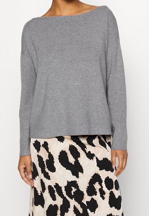 Femme portant un pull gris clair à manches longues et une jupe beige avec un grand imprimé léopard abstrait noir.