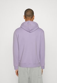 Felpa con cappuccio di colore lavanda realizzata in tessuto morbido, con una vestibilità comoda, polsini e orlo a coste, indossata con pantaloni cargo grigi chiari.