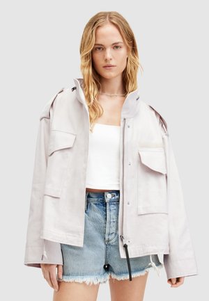 Lichtgrijze oversized jas met een hoge kraag, meerdere zakken aan de voorkant en een ritssluiting, gecombineerd met versleten denimshorts.