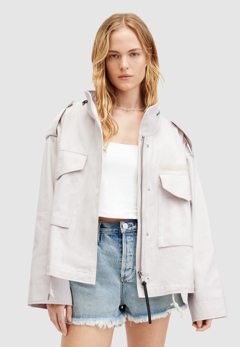 Chaqueta oversized color gris claro con cuello alto, múltiples bolsillos frontales y cierre de cremallera, combinada con pantalones cortos de denim desgastados.