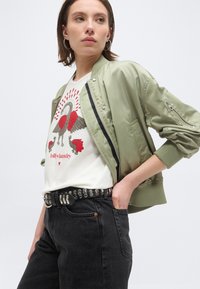 Νεαρή γυναίκα που φοράει ανοιχτοπράσινο μπουφάν τύπου bomber, λευκό γραφικό t-shirt με κόκκινα και καφέ πουλιά, μαύρη ζώνη με τρουκς και σκούρο τζιν.