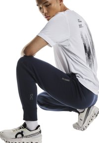 Sportbekleidung: Weißes T-Shirt mit schwarzem Aufdruck, dunkelblaue Jogginghosen und weiße Laufschuhe mit schwarzen Akzenten. Glatter Stoff und eng anliegendes Design, atmungsaktiv.