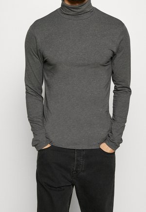 Mann trägt engen dunkelgrauen Langarm-Rollkragenpullover und schwarze Hose mit Händen in den Taschen vor einfachem Hintergrund.