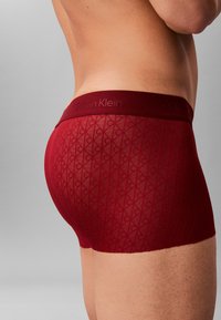 Czerwone bokserki Calvin Klein z teksturowanym wzorem w diamenty i markowym pasem. Materiał wydaje się gładki i dopasowany.