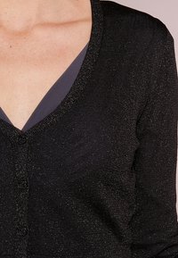 Svart v-ringad kofta i texturerat material med subtil lyster, med knäppningar fram och en kontrasterande mörkgrå innerfoder.