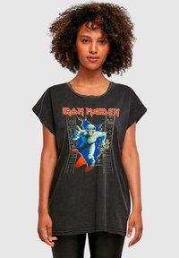 Must on suur must t-särk, millel on graafiline trükk muumiast ja tekst "Iron Maiden" punases värvis, vanutatud tekstuuriga ja rullitud varrukatega.