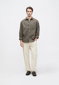 Joven de pie con una mano en el bolsillo, vestido con una camisa marrón abotonada, pantalones beige holgados y zapatos marrones oscuros sobre fondo blanco.