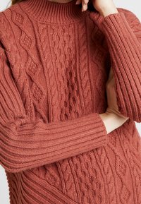 Maglione chunky in terracotta con texture a coste e motivi a treccia. Collo alto e maniche lunghe con una vestibilità rilassata.