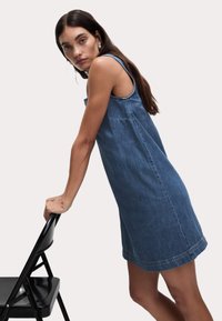 Sukienka dżinsowa w średnim odcieniu niebieskiego, bez rękawów, o luźnym kroju, z widocznymi przeszyciami i linią dołu sięgającą ponad kolano.