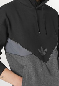 Person som har på sig en färgblockerad svart, grå och mörkgrå Adidas hoodie med framficka och huva, hand delvis i fickan.