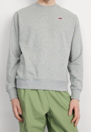 Person trägt ein hellgraues Sweatshirt mit Rundhalsausschnitt und kleinem roten Turnschuh-Aufnäher auf der Brust sowie locker sitzende hellgrüne Hose.