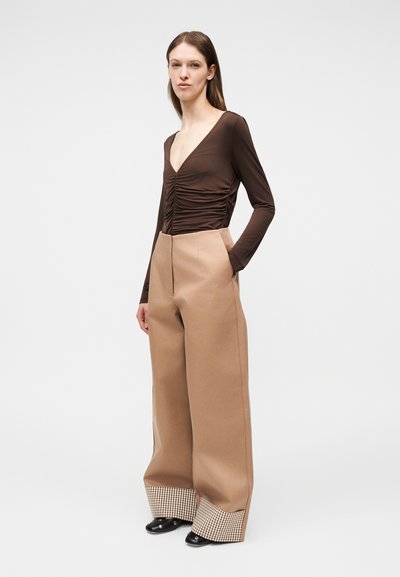 Γυναίκα με μακριά καστανά μαλλιά φοράει καφέ τοπ με μακριά μανίκια και πιέτες και φαρδιά μπεζ παντελόνια με μανσέτες με καρό σχέδιο, στέκεται με τα χέρια στις τσέπες.