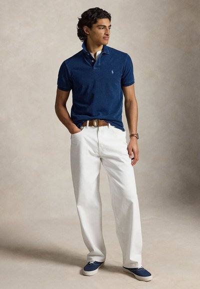 Man standing with hands in pockets, wearing a navy polo shirt, white pants, brown belt, and blue sneakers, looking to the side.