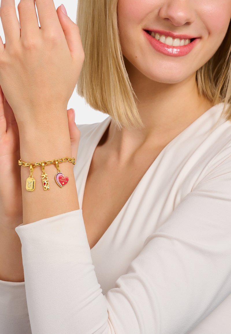 Goldene Armbandkette mit drei Charms: einem rechteckigen goldenen Charm, einem pinken Herzen und einem roten und goldenen Akzent-Charm, vor einem hellen Hintergrund.