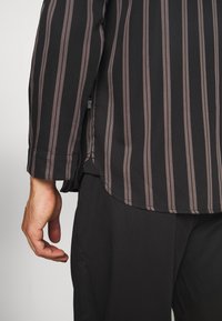 Schwarzes Langarmshirt mit braunen vertikalen Streifen. Hat einen gerundeten Saum und knöpfbare Manschetten. Nahaufnahme einer hand, die auf schwarzen Hosen ruht.