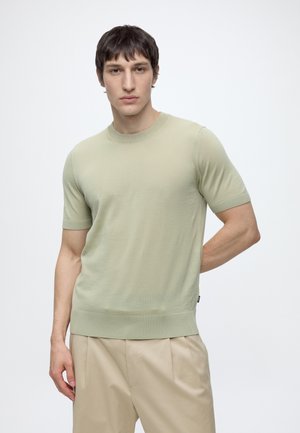 Jeune homme aux cheveux courts et foncés portant un pull vert clair à manches courtes et un pantalon beige, debout devant un fond blanc uni.