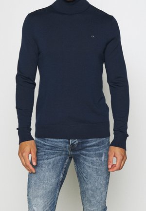 Homme portant un pull à col roulé bleu marine avec un petit logo sur la poitrine et un jean bleu clair délavé, debout devant un fond uni.