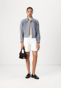 Giacca bomber grigia sopra una maglietta beige, pantaloni bianchi e mocassini neri. Tiene una piccola borsa nera con dettagli bianchi. Outfit semplice e casual.
