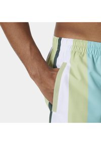 Ręka spoczywa w kieszeni elastycznych spodenek z poziomymi paskami w kolorach zielonym, białym i jasnoniebieskim na jednolitym tle.