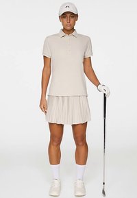 Beżowa koszulka polo z trzema guzikami, plisowana spódnica, biała czapka, białe rękawiczki golfowe oraz kij golfowy. Gładkie materiały, sportowy design i dopasowany krój.