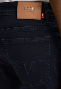 Granatowe dżinsy z denimu z brązową skórzaną metką z napisem "JOOP! JEANS." Posiadają tylną kieszeń z kontrastowym wzorem przeszycia.