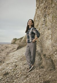 Wolliger navy-blauer Pullover mit beigem Karomuster, kombiniert mit hellgrauen, locker geschnittenen Hosen und braunen Sneakers, vor einem felsigen Strandhintergrund.