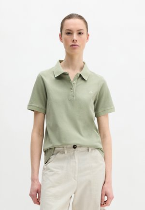 Frau trägt ein hellgrünes kurzärmeliges Poloshirt und beige Hosen, steht vor einem schlichten weißen Hintergrund.