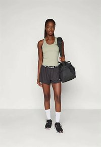Young woman in athletic wear holding a black gym bag, wearing a tank top, shorts, white socks, and black sneakers, standing against a plain background.