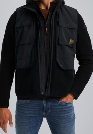 Homme portant un gilet noir zippé avec deux grandes poches avant sur un pull noir et un jean bleu, main dans la poche.