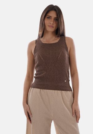 Giovane donna che indossa un top marrone senza maniche in maglia a coste e pantaloni beige larghi, in piedi contro uno sfondo chiaro e uniforme.