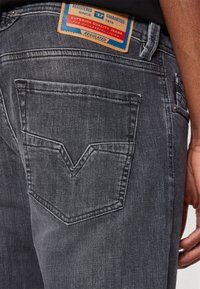 Tmavě šedé džínové kalhoty s výrazným V-švovým prošíváním na zadních kapsách a hnědou etiketu s modrým a červeným nápisem. Viditelný detail texturované látky.