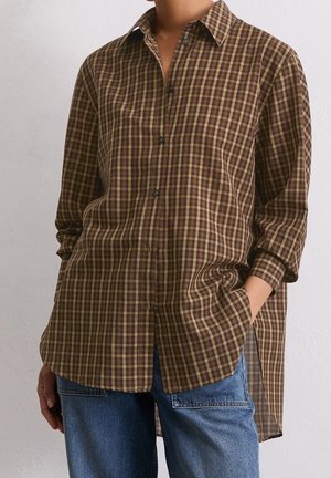 Personne portant une chemise à carreaux marron et jaune à manches longues boutonnée, avec un jean bleu, une main dans la poche, debout devant un fond uni.