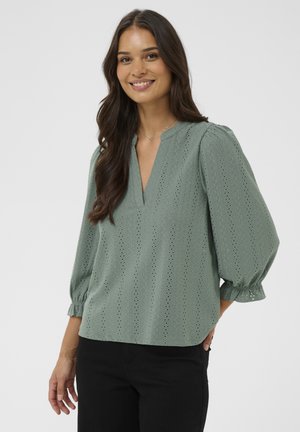 Blouse verte texturée avec détails en œillets, col en V et manches bouffantes. Associée à un pantalon foncé avec des poignets froncés.